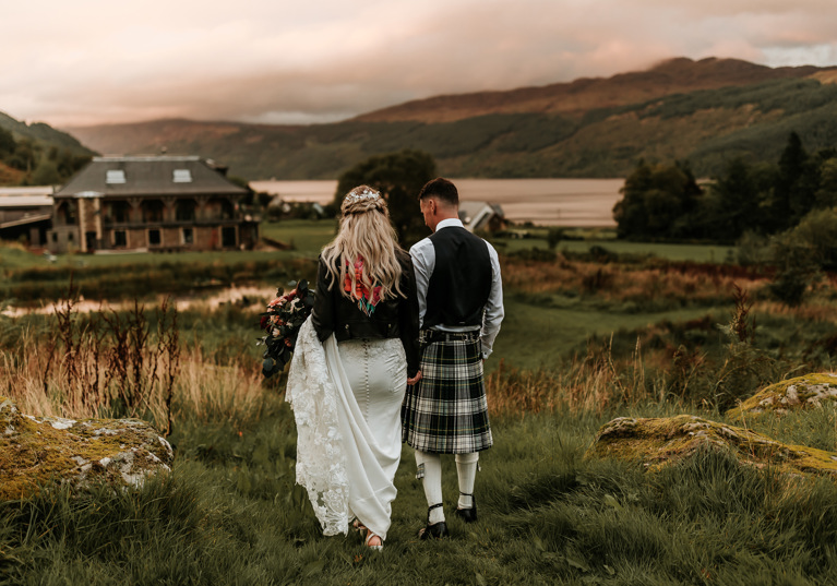 Bride wearing leather jacket and groom wearing kilt walk through grass